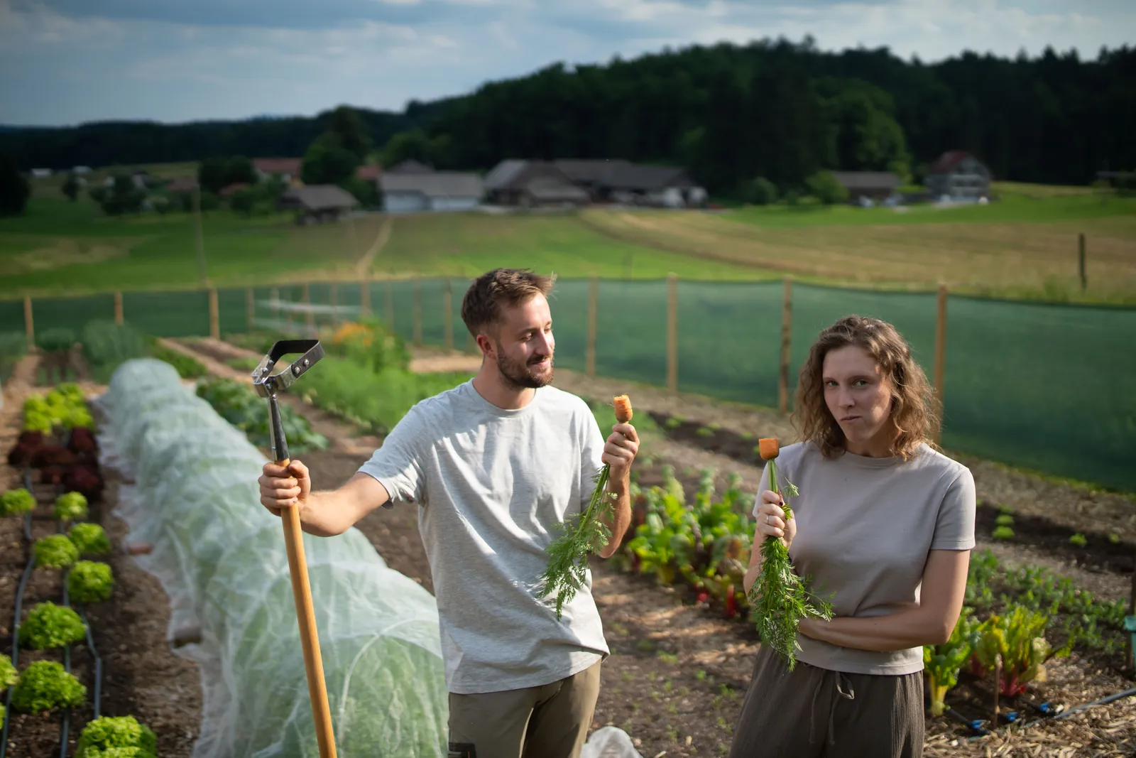 Jure and Ajda in the garden holding freshly pulled carrots, a broadfork and garden beds beside them.